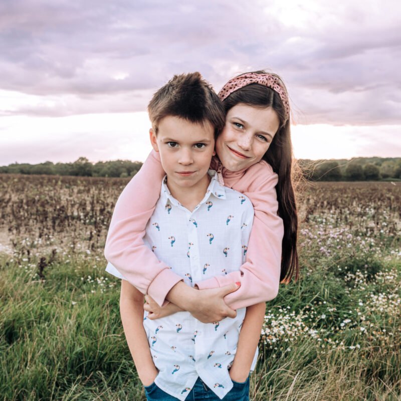 An older sister, hugging her brother from behind whilst they look into the camera, standing in a West Sussex field at sunset