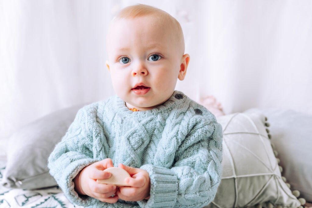 Sitting baby in the stuido, wearing a chunky mint-green jumper, looking into the camera with big blue eyes, with cushions behind him.