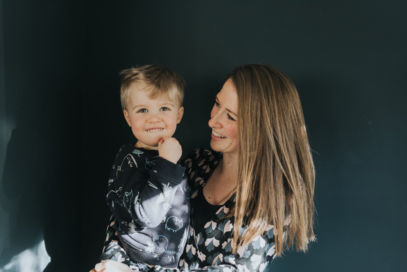 A photography of a mum holding her 2 year old son against a dark blue wall.