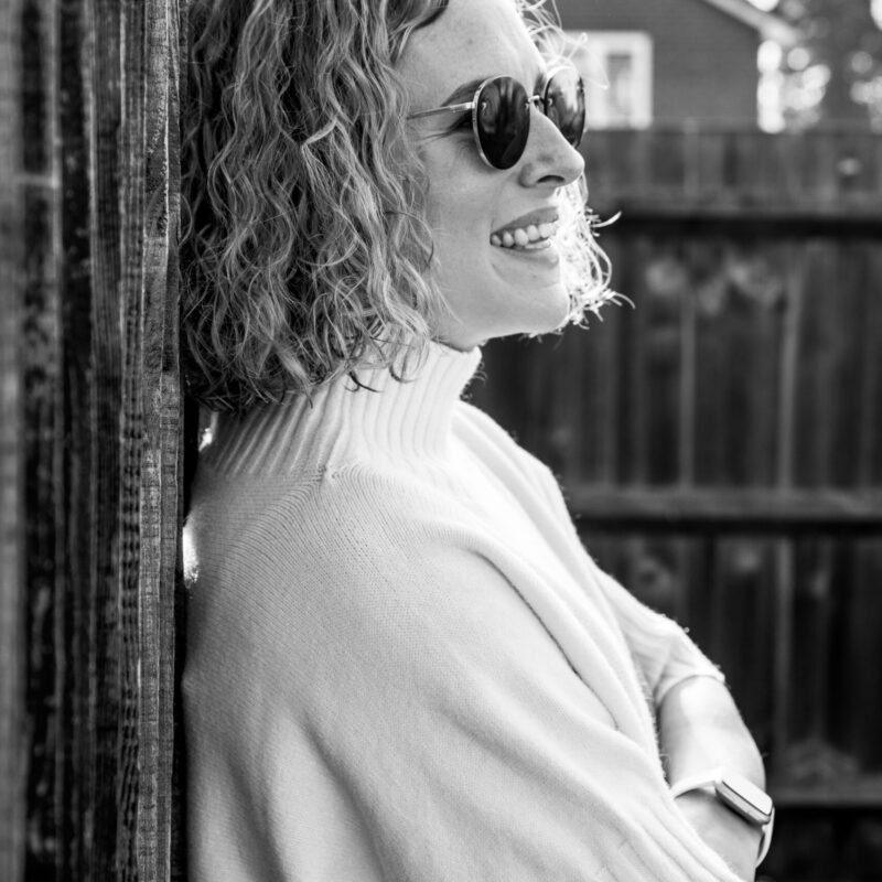 A black and white portrait of a careers coach standing against a fence, with sunglasses on and her arms crossed looking out, smile on her face, feeling relaxed.