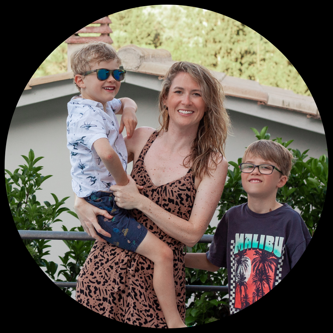 A mum and her two sons on holiday in Greece, smiling at the camera and holding her 5 year old on her hip.