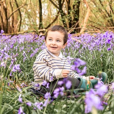 A little boy sat amongst the bluebells in a West Sussex woodland in his green wellies and strip top, smiling.